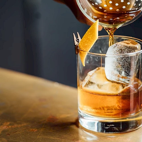 A whiskey cocktail being poured into a rocks glass with a citrus twist garnish and a large ice cube, on a bar counter.