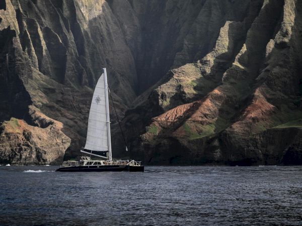 A sailboat glides near rugged, dark cliffs with light on the rock faces, water calm, creating a striking coastal scene.