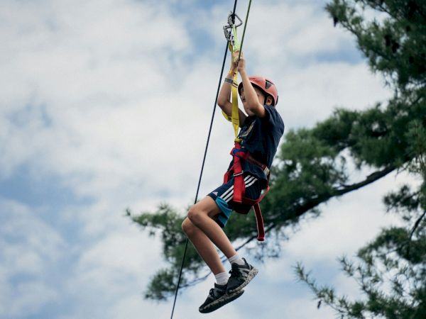 A child or teen zip-lining/climbing a rope between trees, wearing a helmet and harness against a blue sky.