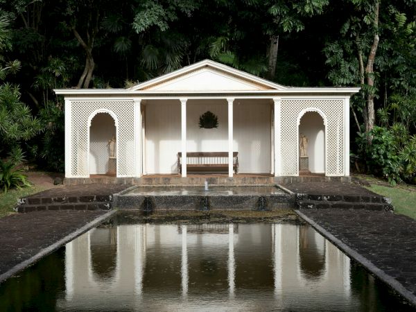 A small, white neoclassical pavilion with arches and benches, set beside a reflecting pool in a wooded garden.