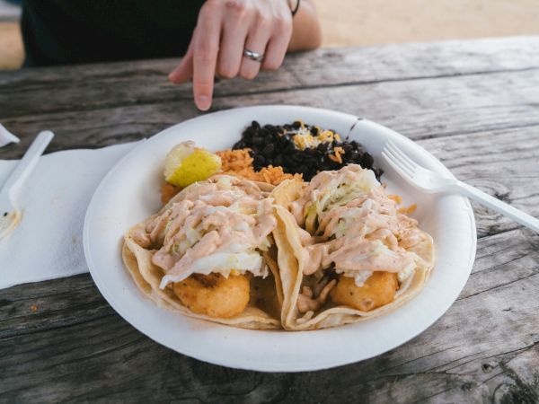 A plate with dumplings in sauce, rice and seaweed, plus yellow pickle and a potato croquette; someone&rsquo;s hand with a ring nearby, on a wooden table.