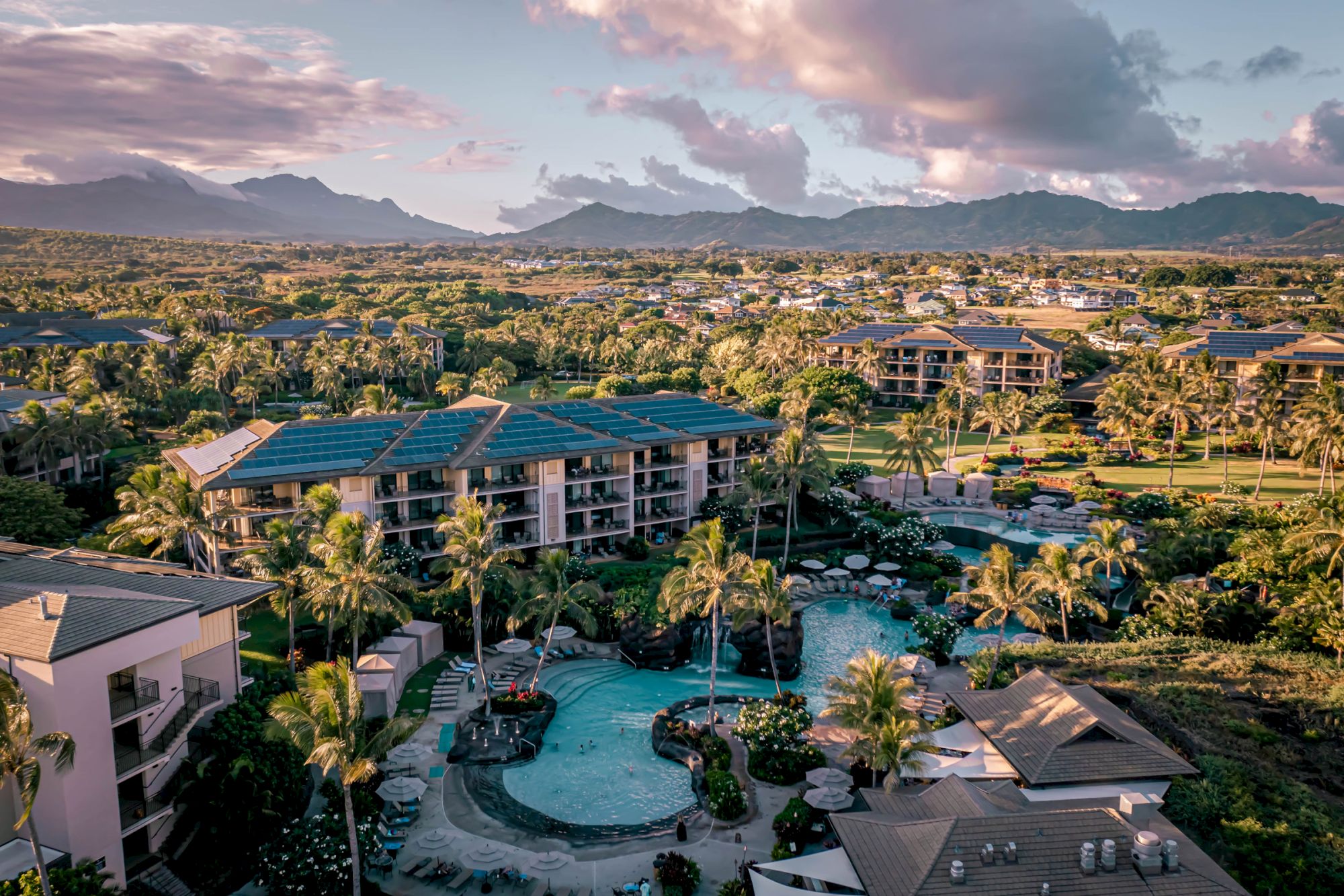 A resort complex with multiple buildings, palm trees, and a winding pool area, set against a distant mountain range at sunset.