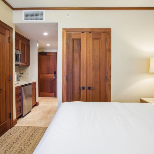 A bedroom with wooden doors, a kitchenette, a bed, teal pillows, and a lamp on a bedside table, creating a cozy atmosphere.