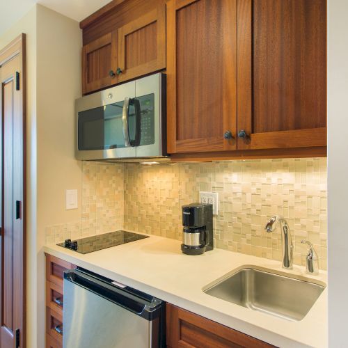 A compact kitchen with wooden cabinets, a microwave, a small sink, a coffee maker, and a mini-fridge below a light-tiled backsplash.