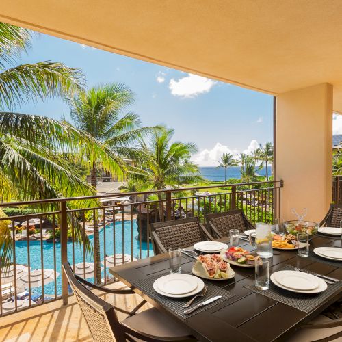 A balcony view with a table set for dining overlooks a pool, palm trees, and a blue sky.