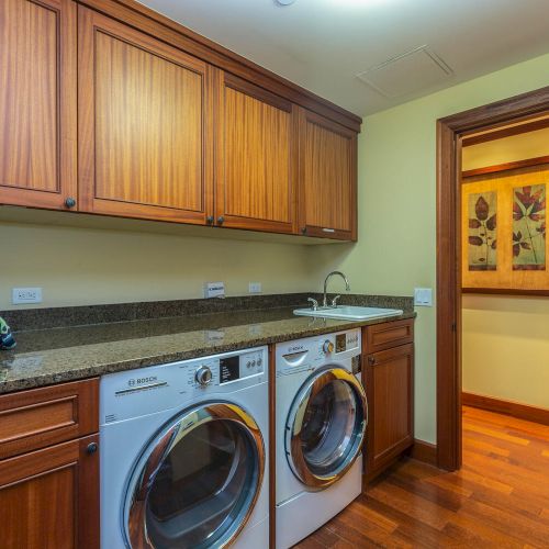 A laundry room with a washer and dryer, wooden cabinets, folded towels, a sink, and a hallway with framed artwork on the wall.