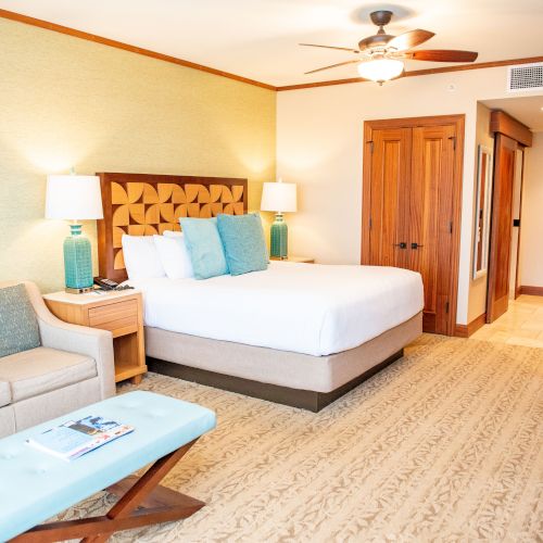 A hotel room features a bed, sofa, and decor, with warm lighting and a ceiling fan, creating an inviting atmosphere.