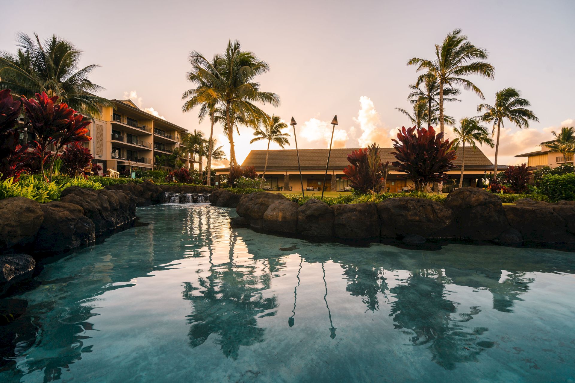 A tropical resort scene with a stone-lined pool, waterfalls, palm trees, colorful plants, and hotel buildings at sunset.
