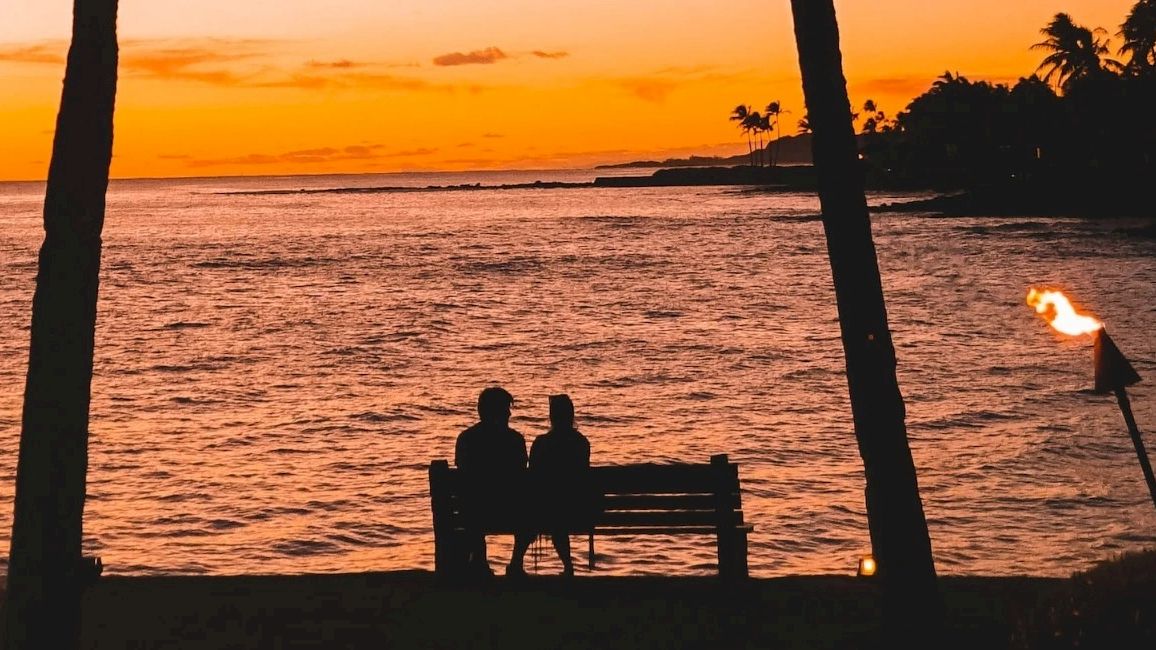 Silhouetted couple sit at a beach bench as palm trees, torches, and a glowing sunset paint the ocean.