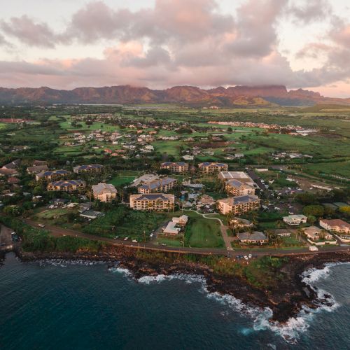 Coastal coastline with rocky shore, clusters of houses and green fields inland, calm ocean water, and a distant mountain range under a partly cloudy sky.