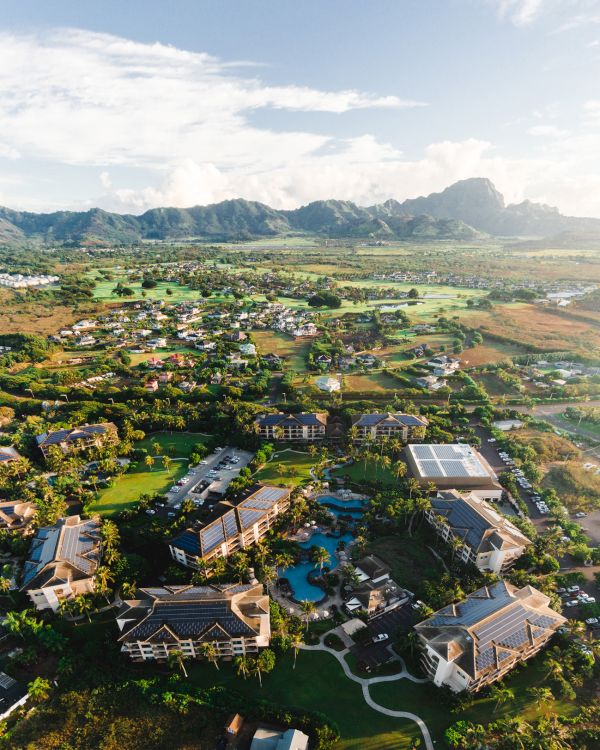 Aerial view of a lush resort town with buildings around a central pool, green lawns, and distant mountains under a bright sky.