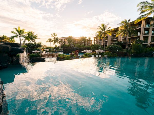 Resort resort-style pool with turquoise water, palm trees, sunlit buildings, and lounge areas reflecting on the calm surface. End.