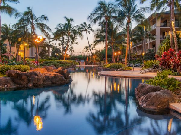 A tropical resort pool glows at dusk, palm trees line the water, lanterns reflect on the calm surface, and resort buildings loom in the background.