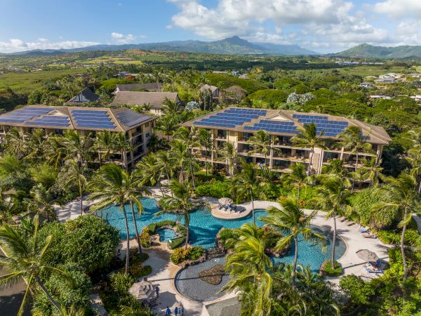A tropical resort with multiple buildings, lush palm trees, and a winding pool area set against a green valley and blue skies.
