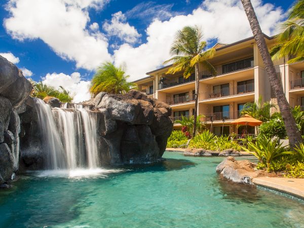 A tropical resort with a cascading waterfall into a turquoise pool, sunlit rocks, palm trees, and a multi-story beige hotel in the background.