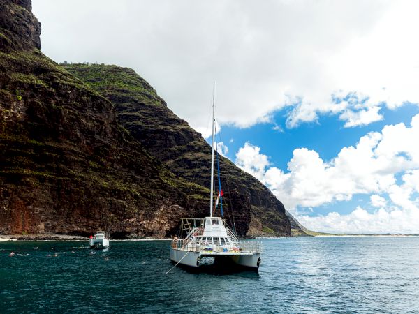 A sailboat is anchored near a rocky coastline with cliffs, clear blue water, and a partly cloudy sky in a scenic seaside setting.