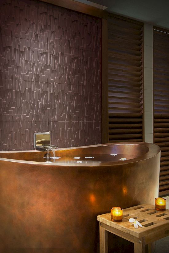 A copper freestanding bathtub in a dim, spa-like room with wooden slat walls, a small stool and candles for a relaxing bath moment.