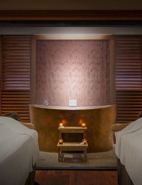 Two massage beds face a wooden-paneled wall with a small lit altar and a bathtub or spa tub in the center, creating a calming, spa-like room.