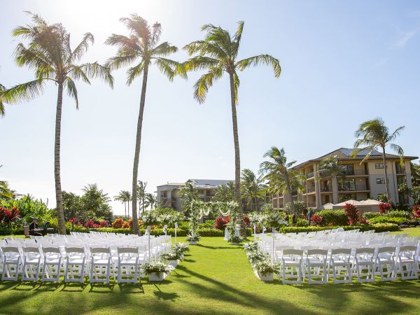 A tropical outdoor wedding setup on a sunny lawn with white chairs, palm trees, and a resort-style building in the background.