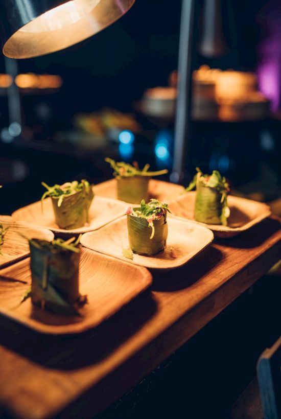 Small, elegantly plated food items, possibly appetizers, garnished with greens, on wooden plates under warm lighting.