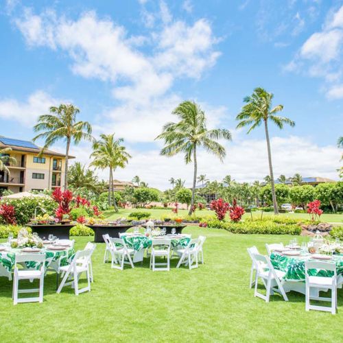 A sunny outdoor lawn setup with round tables and white chairs, turquoise tablecloths, palm trees, and a resort-style building in the background.