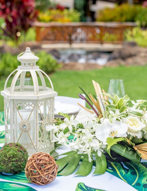 A garden table setup with a white lantern, floral centerpiece, glassware, and tropical-patterned tablecloth in an outdoor yard.