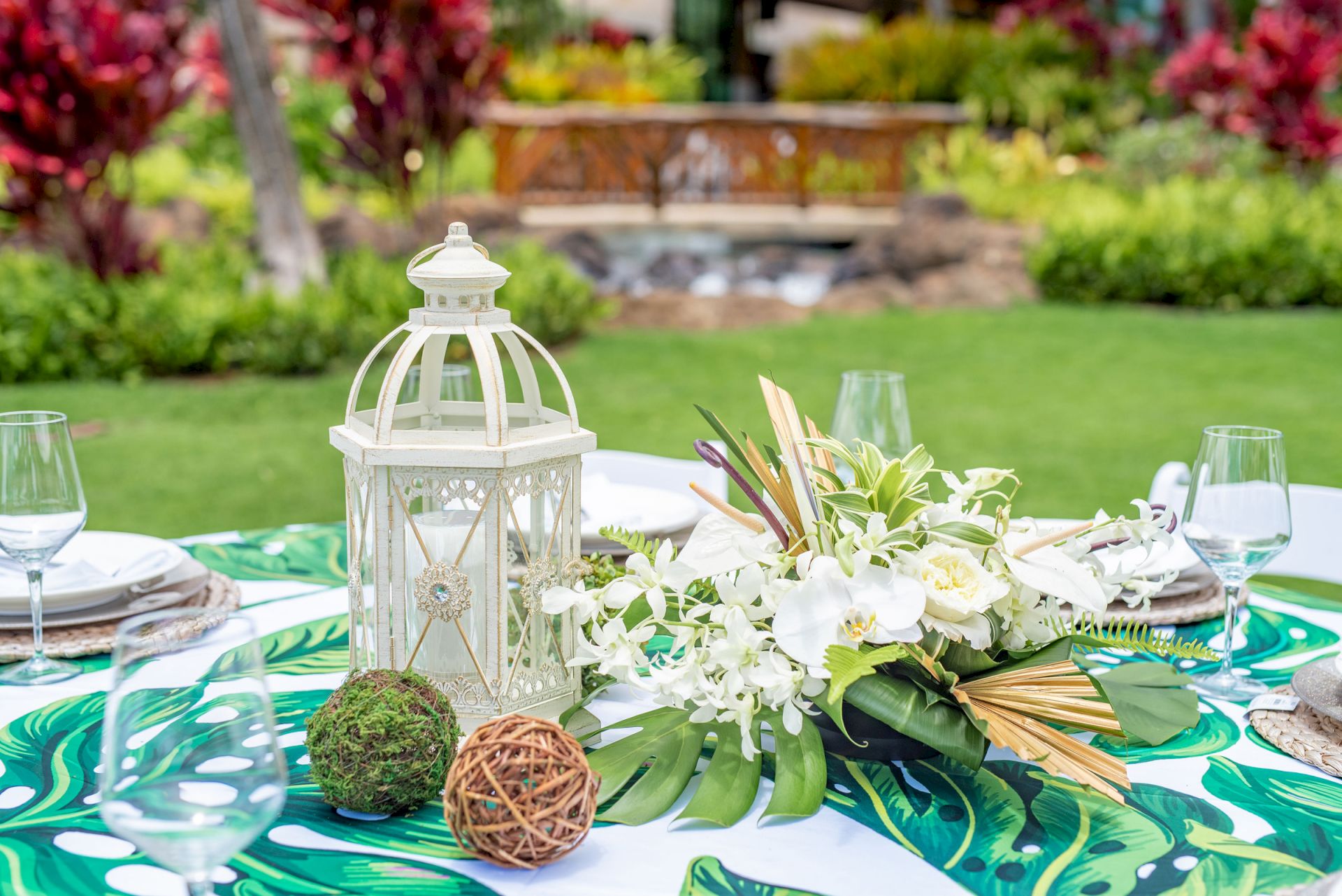 An outdoor spring table setting with a white lantern centerpiece, fresh white flowers, and glasses on a green leaf-patterned cloth.