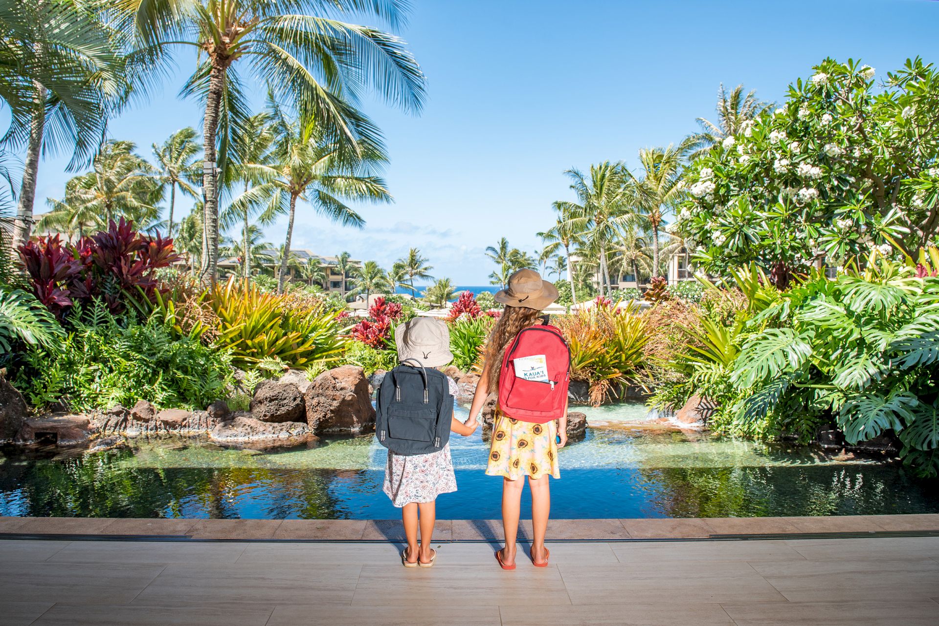Two kids with backpacks stand by a tropical pond amid palms and flowers, enjoying a sunny resort getaway.