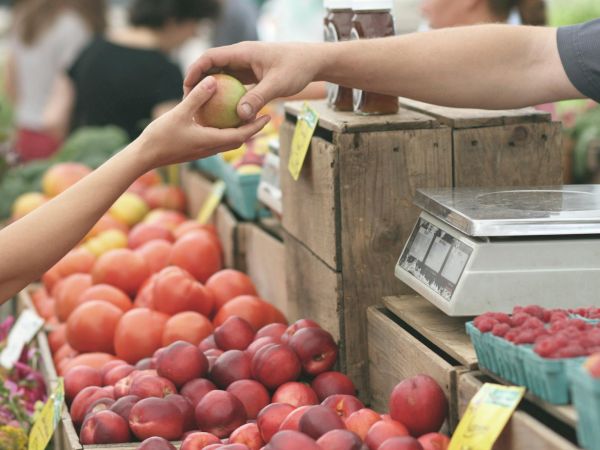 A hand hands a ripe fruit to another across a market stall with peaches, apples, and berries; a scale sits nearby for weighing produce.