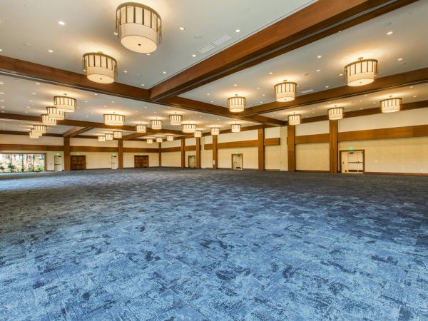 A spacious, empty banquet hall with blue carpet, warm wood beams, and round hanging lights across a long, well-lit room.