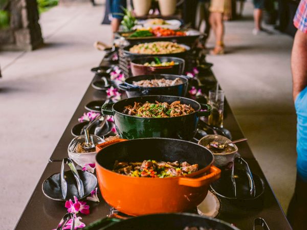 A long buffet line of colorful dishes in pots, guests serving themselves under a covered outdoor area, flowers decorating the table.