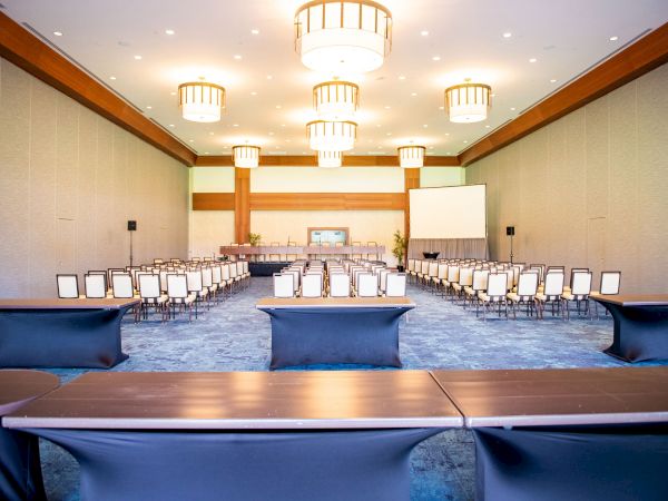 A conference room set up for a large meeting, rows of chairs facing a stage with a projector screen, and round chandeliers overhead.