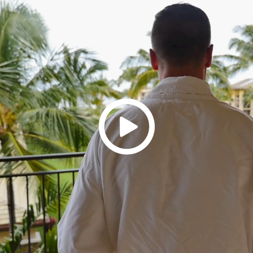 Two people in white robes stand on a balcony with palm trees and tropical scenery in the background.