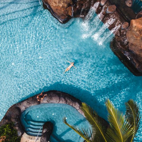 A person floats in a tropical pool surrounded by rocks and waterfalls, with clear blue water and lush greenery around.