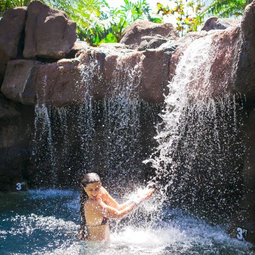A person stands under a waterfall in a pool surrounded by rocks and tropical greenery.
