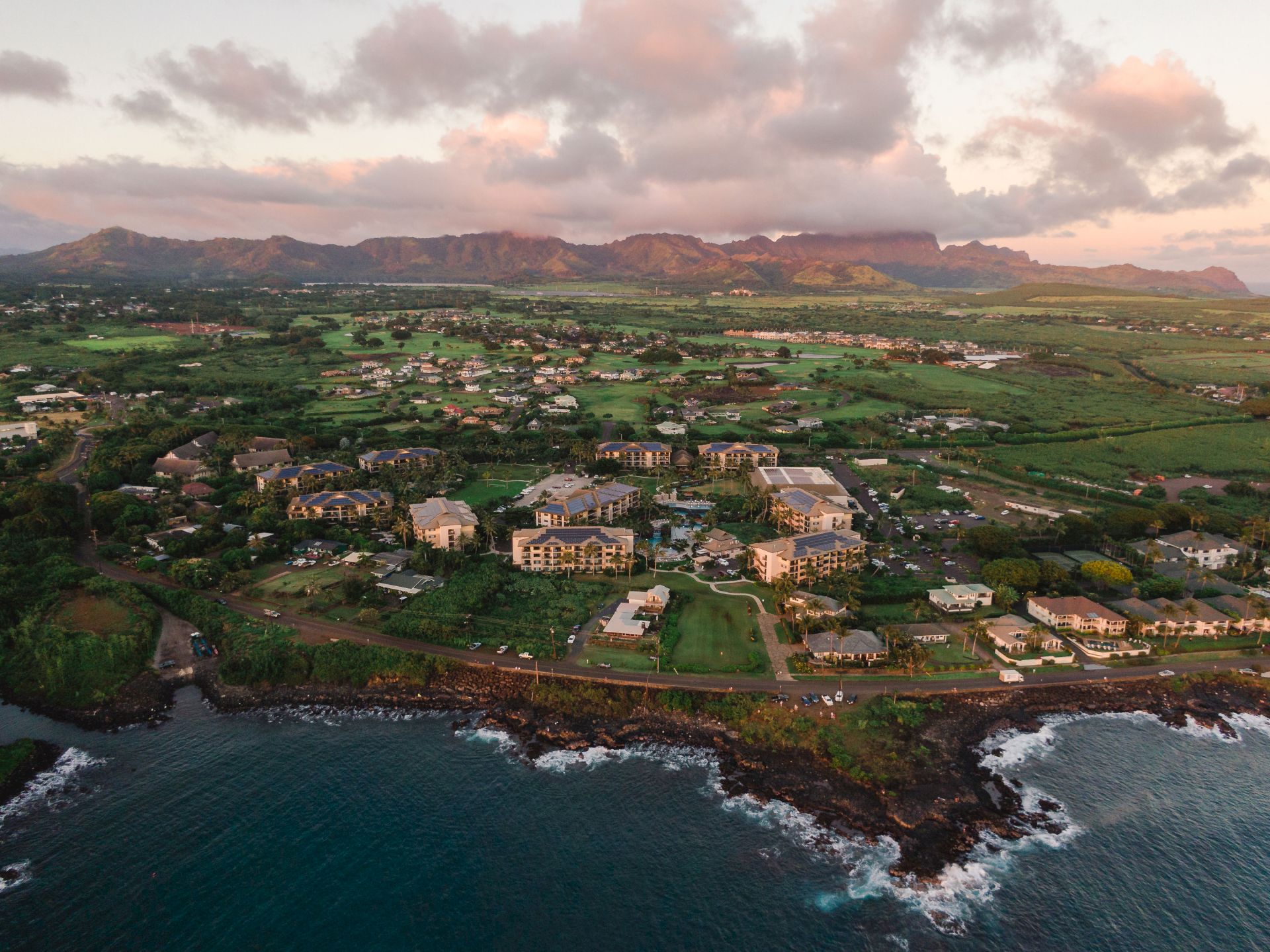 Aerial view of a coastal town with buildings, greenery, and mountains in the background, under a partly cloudy sky.