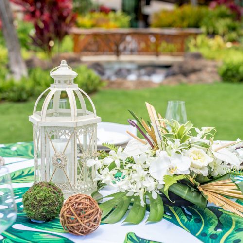 An outdoor table setting with a tropical-themed cloth, decorative lantern, and floral centerpiece, set against a lush garden backdrop.