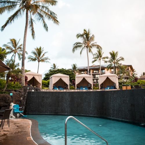 A tropical pool scene with cabanas, palm trees, and resort buildings in the background.