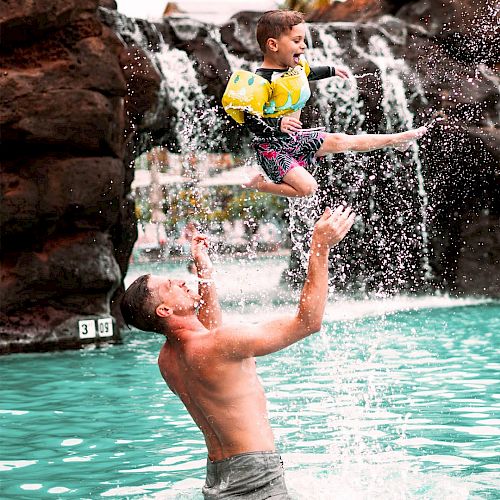 A man is throwing a child in the air at a pool with a waterfall, creating a joyful splash.