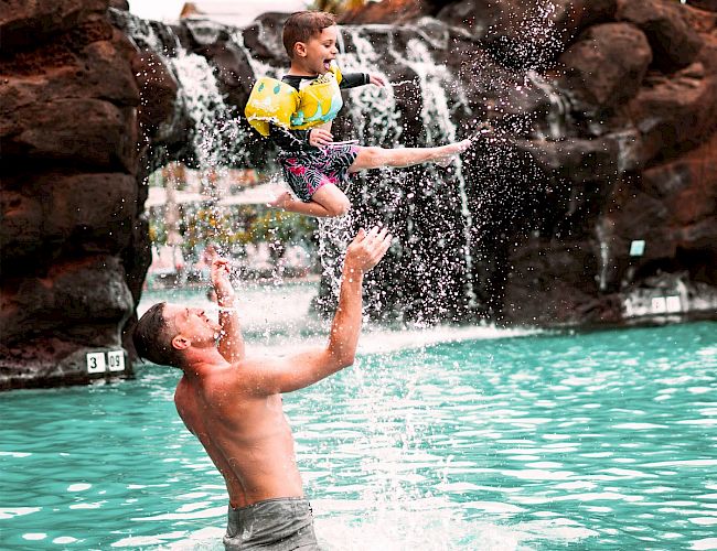 A man is throwing a child in the air at a pool with a waterfall, creating a joyful splash.