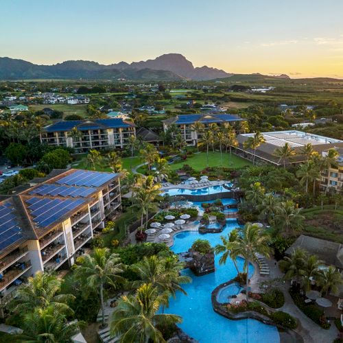 Aerial view of a resort with pools, palm trees, and buildings, surrounded by lush greenery and mountains.