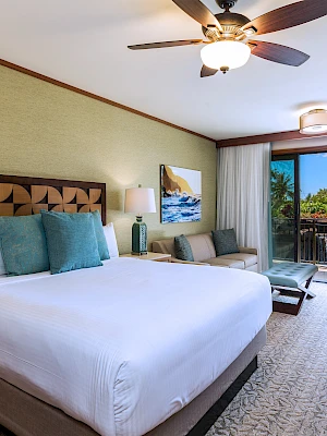 A hotel room with a bed, two lamps, a ceiling fan, and a balcony with a view of greenery and blue skies.