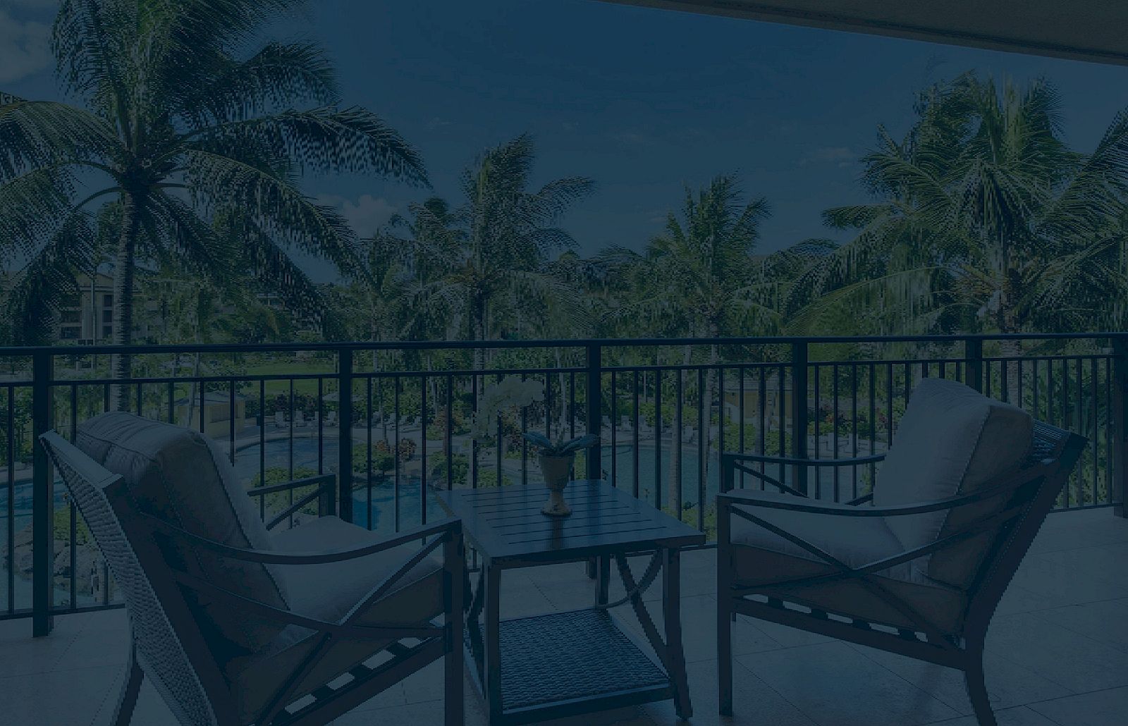 A balcony with two chairs and a small table overlooking a pool and palm trees; a relaxing outdoor seating area at a resort or house.