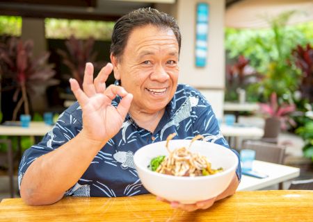 A person smiling, holding a bowl of food, and gesturing "okay" with their hand while sitting at an outdoor table.