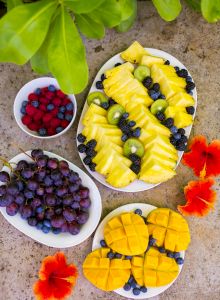 A colorful fruit platter with pineapple, kiwi, blueberries, mango, grapes, and raspberries, accompanied by red flowers on a stone background.
