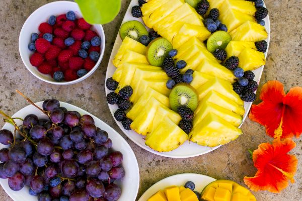 A colorful fruit platter with pineapple, kiwi, blueberries, mango, grapes, and raspberries, accompanied by red flowers on a stone background.