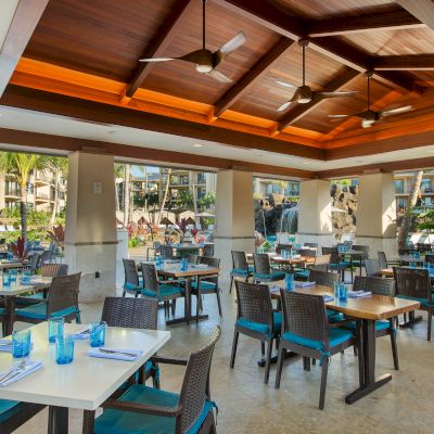 The image shows a bright, spacious restaurant with blue-accented tables, modern wooden ceiling, and large windows overlooking a pool area.