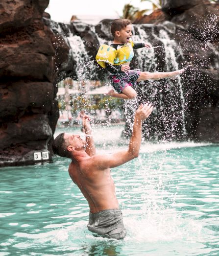 A man in a pool throws a child into the air, with a waterfall and rocks in the background.