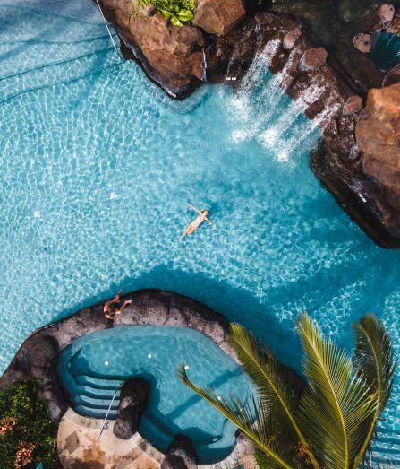Aerial view of a pool with a person swimming, surrounded by rocks and waterfalls, adjacent to a hot tub area with a palm tree nearby.