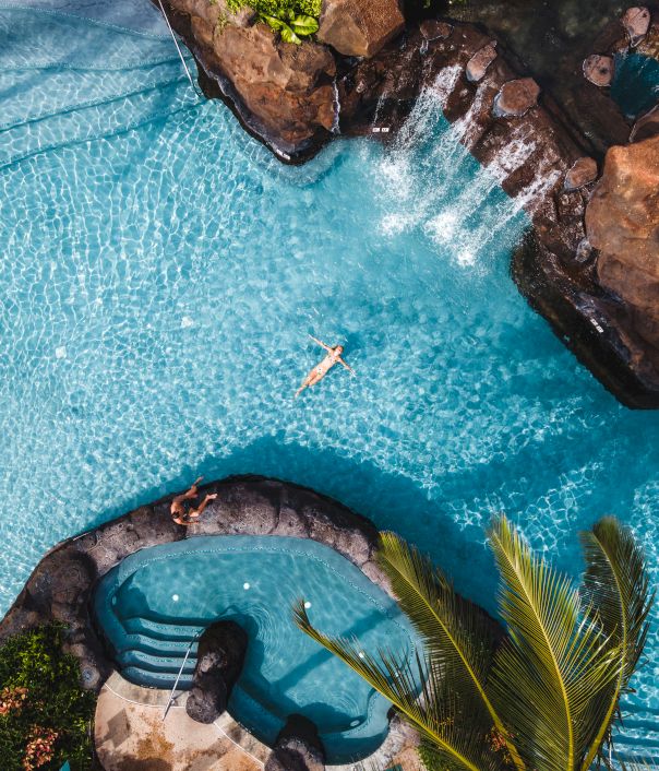 Aerial view of a pool with a person swimming, surrounded by rocks and waterfalls, adjacent to a hot tub area with a palm tree nearby.